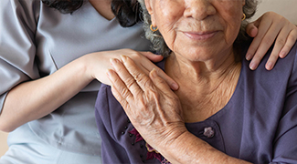 Caregiver standing behind an older woman with her hands on the woman's shoulders, while the older woman smiles and puts her hand one of the caregiver's hand 