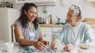 Senior lady having fun building a puzzle at a kitchen table with a caregiver