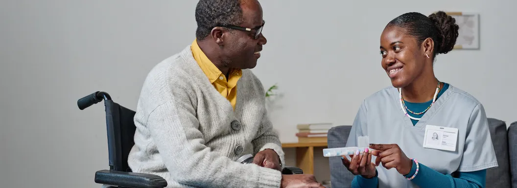 Caregiver showing organizer with pills to man with disability and explaining which pills to take while they sitting in room