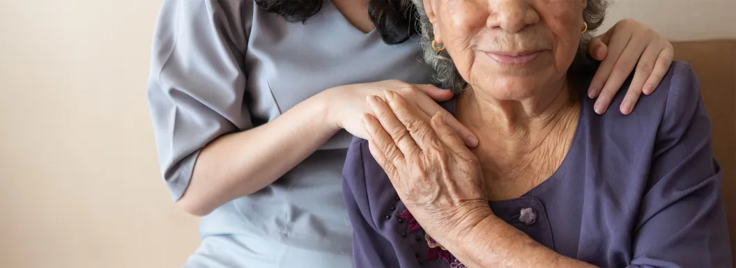 Caregiver standing behind an older woman with her hands on the woman's shoulders, while the older woman smiles and puts her hand one of the caregiver's hand 