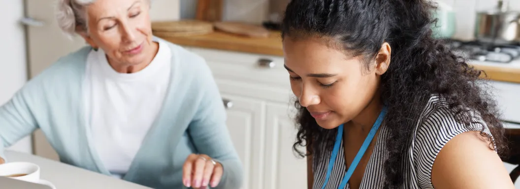 Caregiver helping elderly woman examine a document sitting at kitchen table together in front of laptop