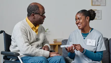 Caregiver showing organizer with pills to man with disability and explaining which pills to take while they sitting in room