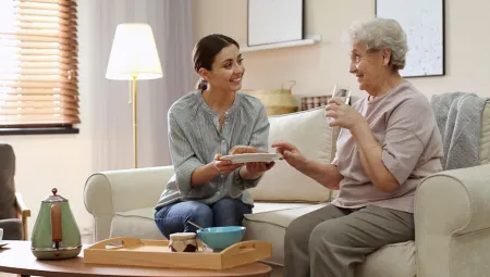 Caregiver holding a plate of food out for senior woman while sitting on a sofa together