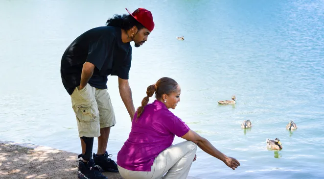 Men bending over next to a woman reaching out to some ducks in a body of water
