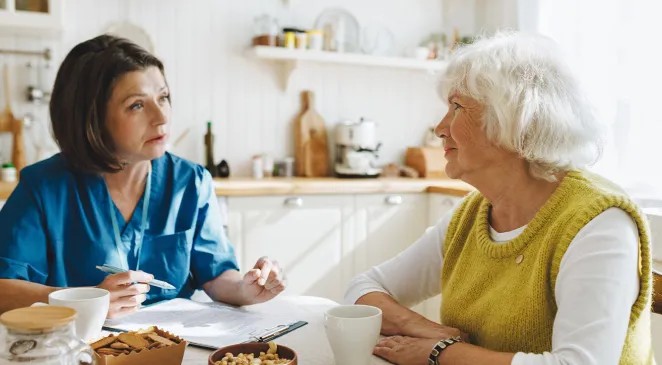 Senior woman answering questions of caregiver sitting at kitchen table drinking tea 