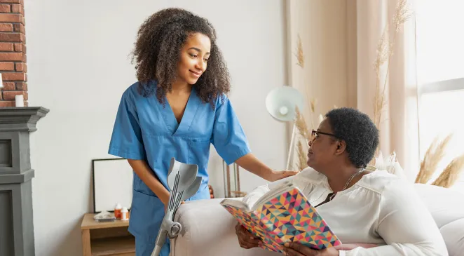 Caregiver with a hand on an elderly woman's shoulder who is sitting down and reading a book while they both smile at each other