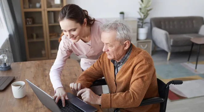 Portrait of senior man in wheelchair using laptop at retirement home with nurse assisting him