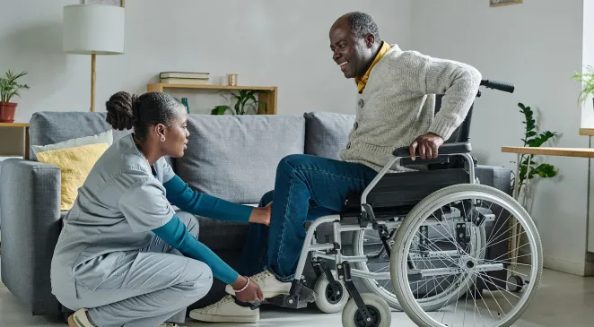 Caregiver crouching while assisting an older man in a wheelchair at home