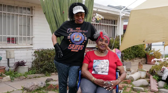 A Home Assist Health caregiver standing next to an elderly woman in wheelchair outside of a house