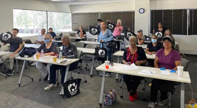 Home Assist Health team members posing for a team photo while sitting at rows of tables for a training with matching black Home Assist Health branded tote bags