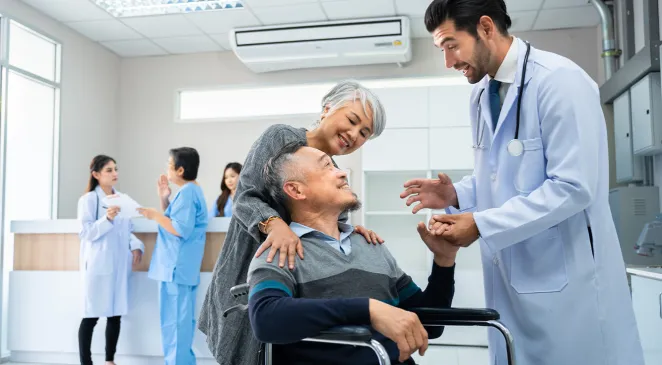 Doctor standing holding hand while talk to a happy senior male patient sitting on wheelchair pushed by a caregiver at a hospital