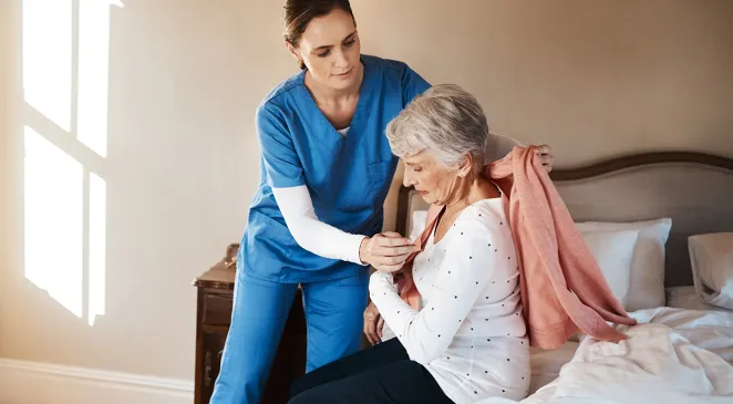 Caregiver assisting senior woman in getting dressed while she sits on her bed