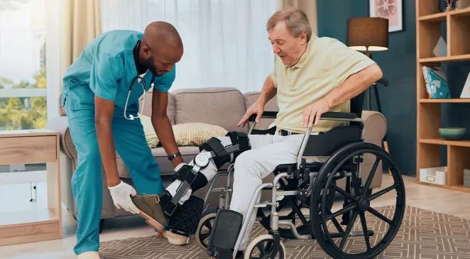 Caregiver assisting an elderly man with a leg brace in a wheelchair put his shoes on
