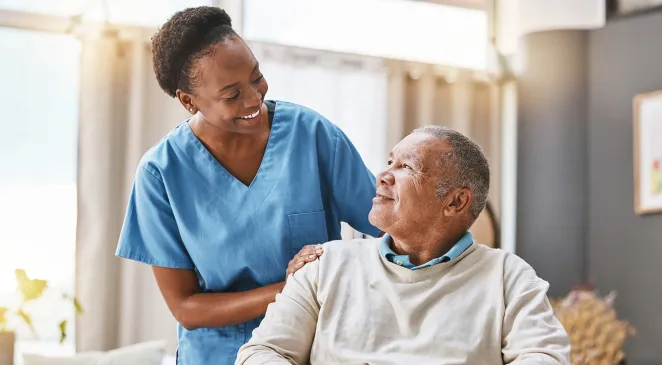 Caregiver in scrubs with her hand on the shoulder of an older man sitting while they smile and look at each other