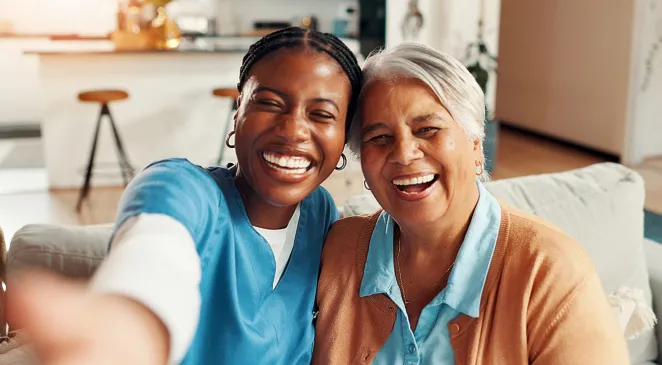 Selfie of smiling nurse with old woman on sofa in living room 
