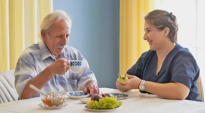 Caregiver cutting an apple and a senior man sipping on a cup of coffee while they sit together at dining table