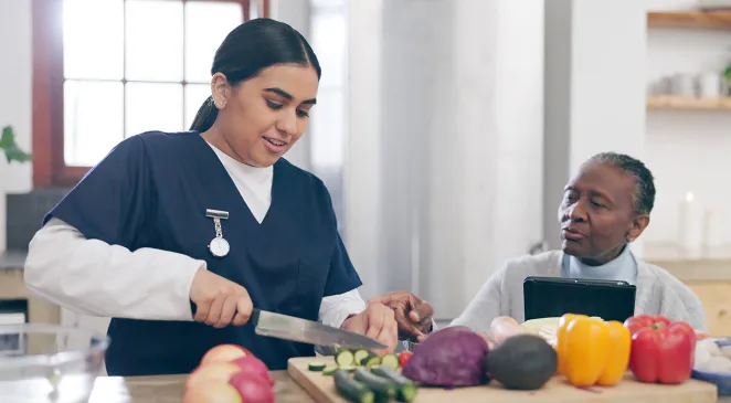 Nurse helping elderly person chop vegetables in a kitchen