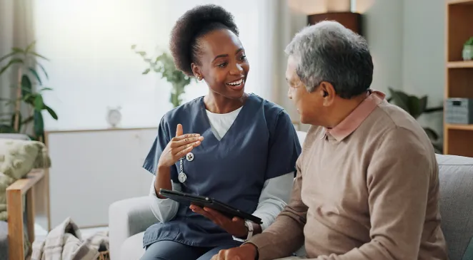 Caregiver explaining something to a senior man while holding a tablet and sitting on a sofa