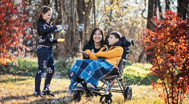 Mother smiling next to a disabled young boy in wheelchair and a young girl smiling while blowing bubble outside