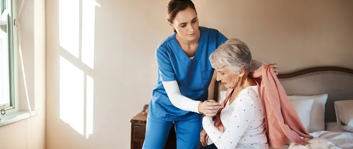 Caregiver helping an elderly woman get dressed while sitting on a bed