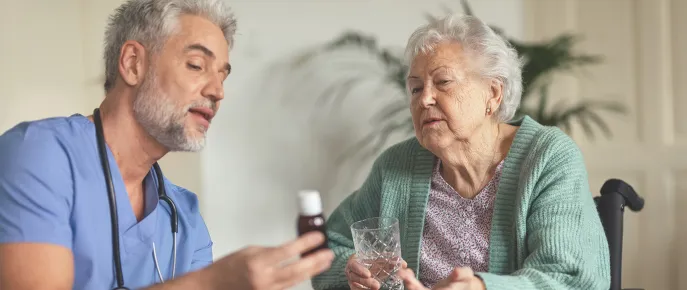 Caregiver showing medicine to an elderly woman holding a glass of water