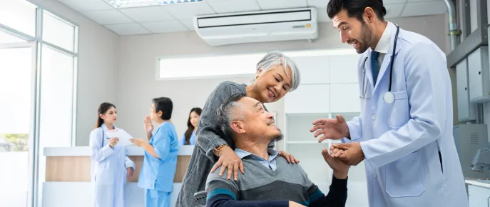 Doctor standing holding hand while talk to a happy senior male patient sitting on wheelchair pushed by a caregiver at a hospital